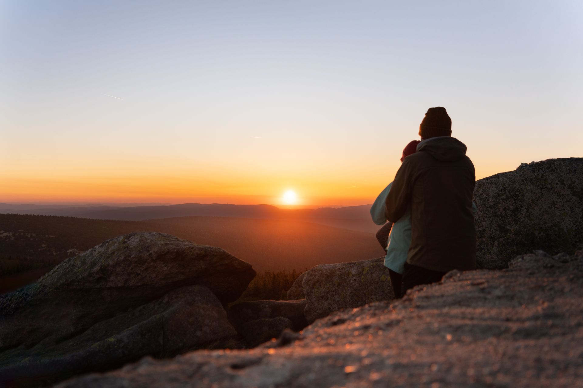 Two people sit on a rocky mountain peak looking out at a sunset over distant hills.