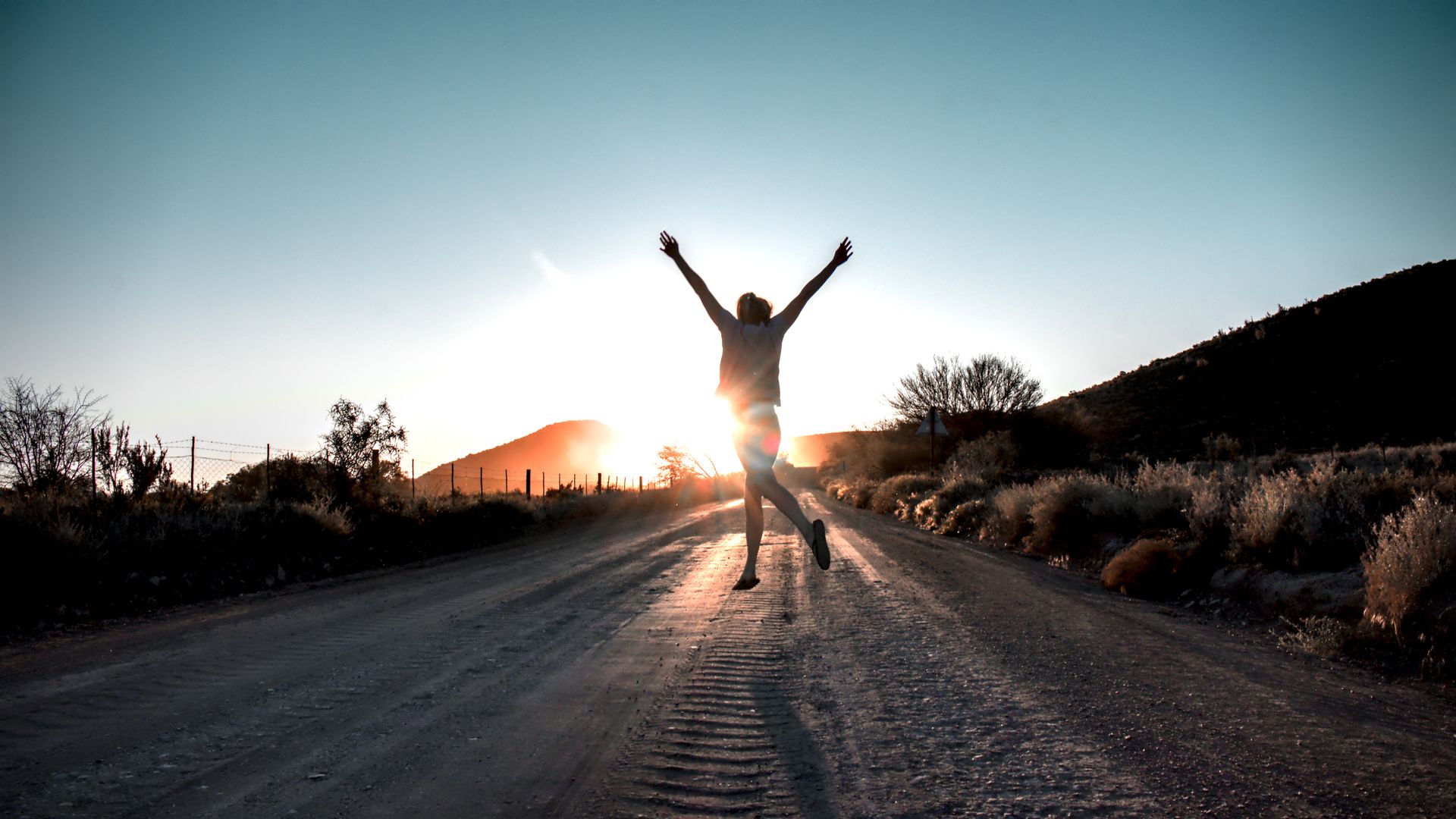 A person jumps for joy in the middle of a dirt road toward a bright sunset.