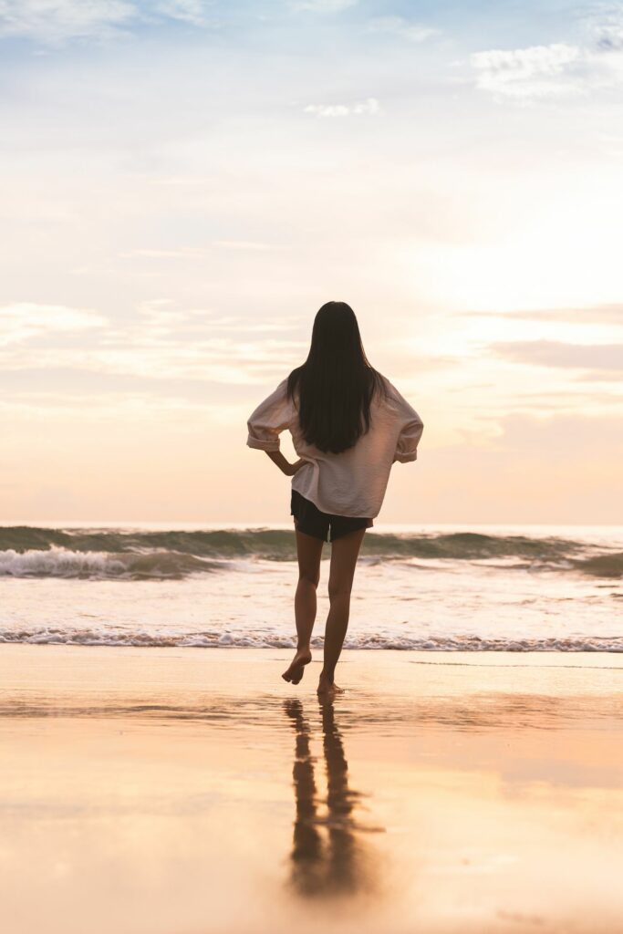 A person with long dark hair stands with hands on hips, looking out at the ocean during sunset.