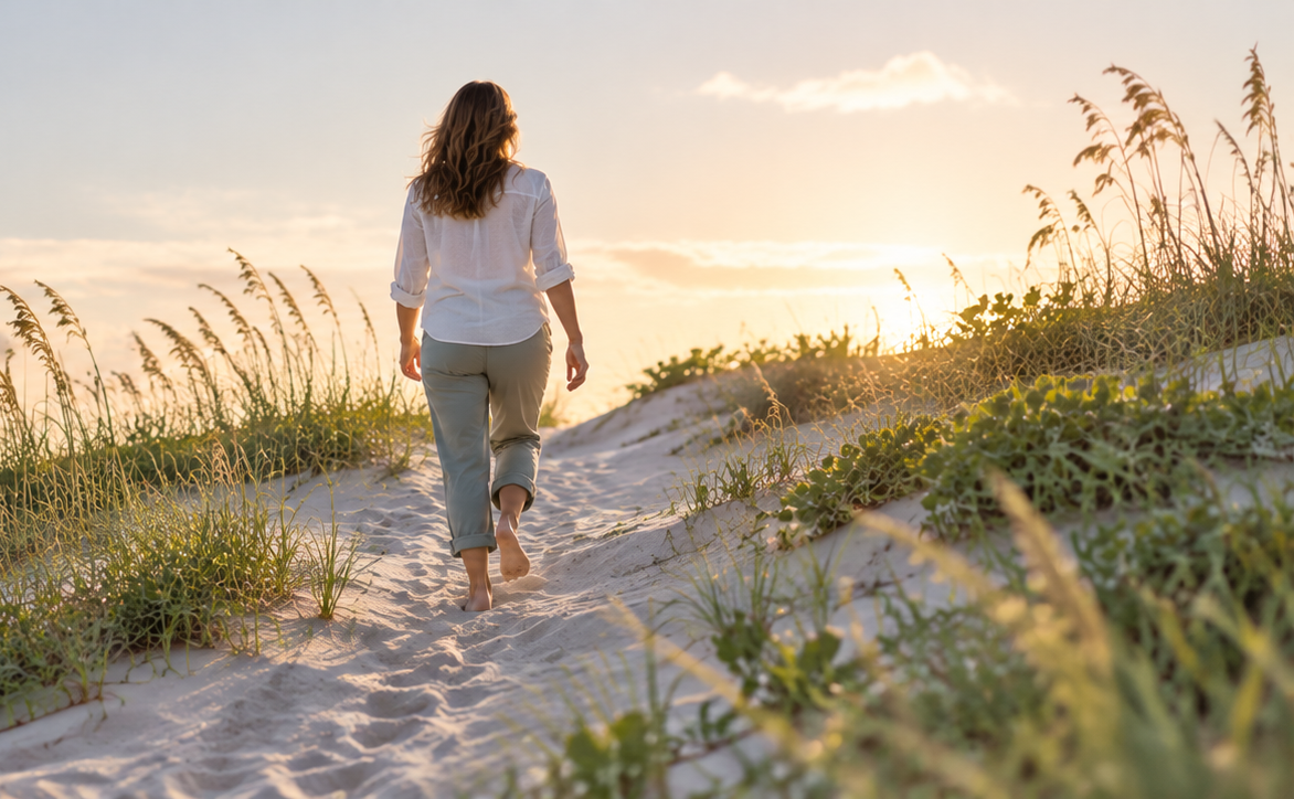 A woman walks barefoot along a sandy path through grassy dunes toward the sunset.